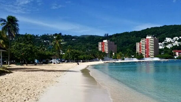 A beach with clear blue water, sandy shore, and buildings in the background.