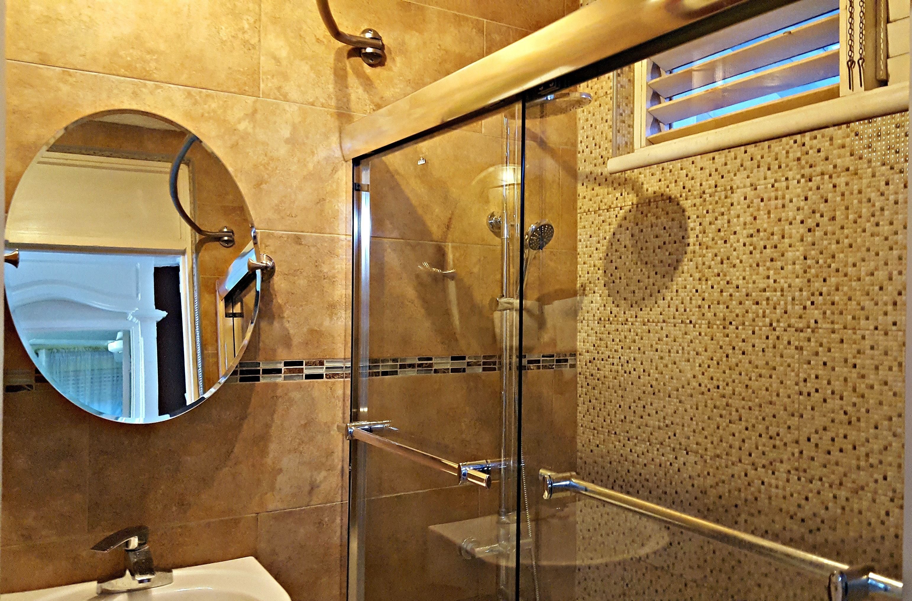 A modern bathroom with a glass shower enclosure, a round mirror, and a sink.