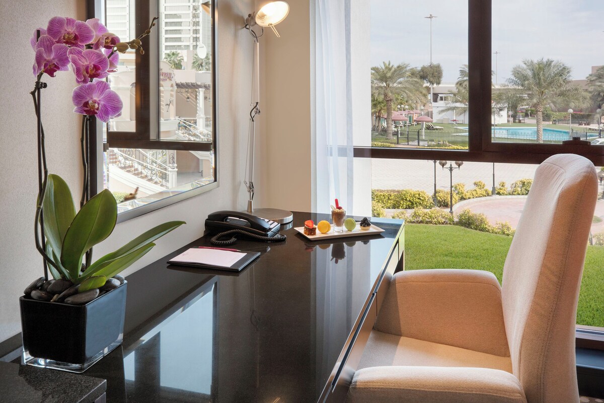 Habitación de hotel con escritorio, una silla, una planta en maceta, teléfono y ventana con vistas a una piscina y palmeras.