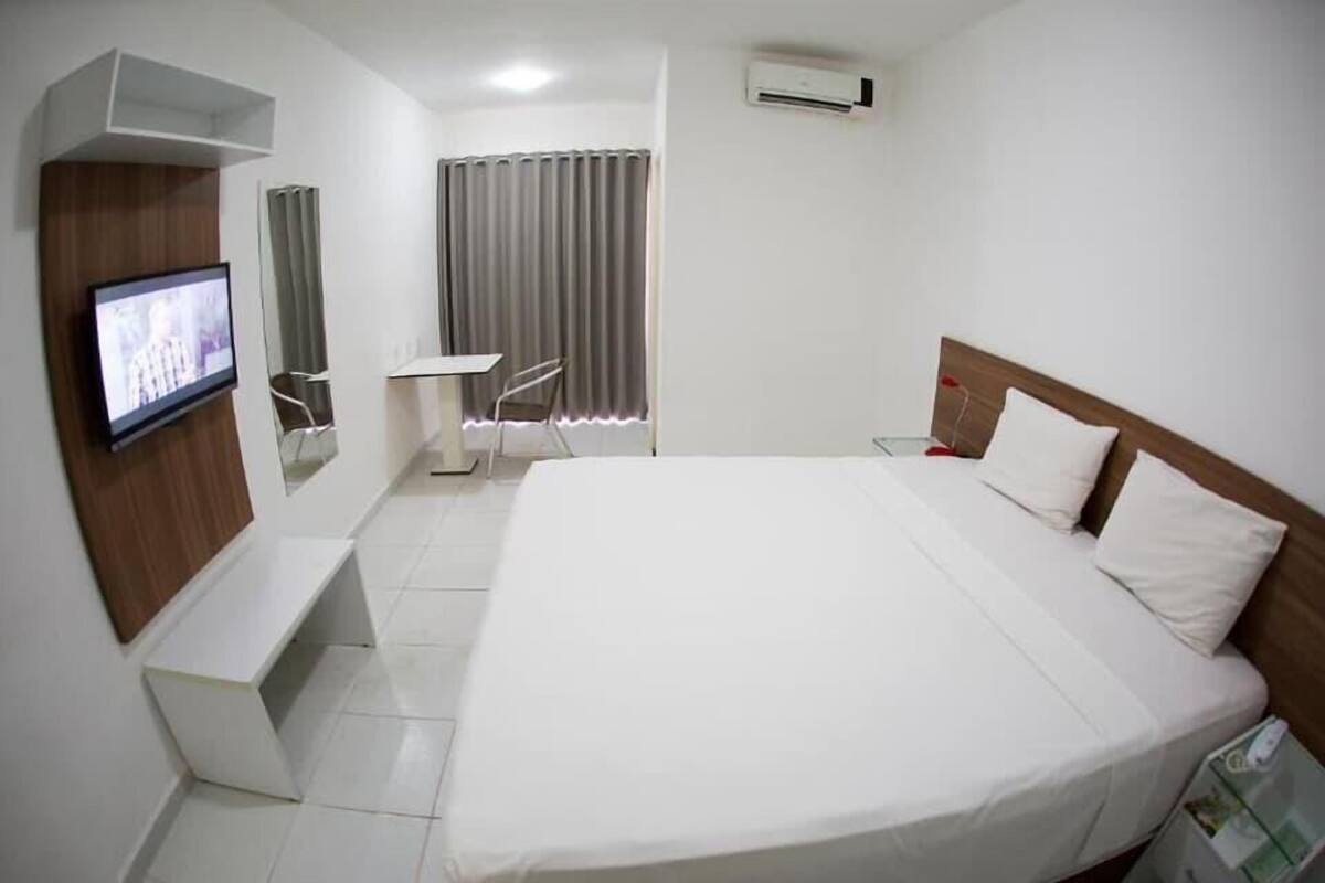 Une chambre d’hôtel comprenant un lit, une télévision, une petite table, une chaise, une fenêtre avec des rideaux et un appareil de climatisation.