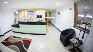 A reception area with a counter, a black chair, a magazine rack, and a tiled floor. - Rede Andrade Araras (Aracaju)