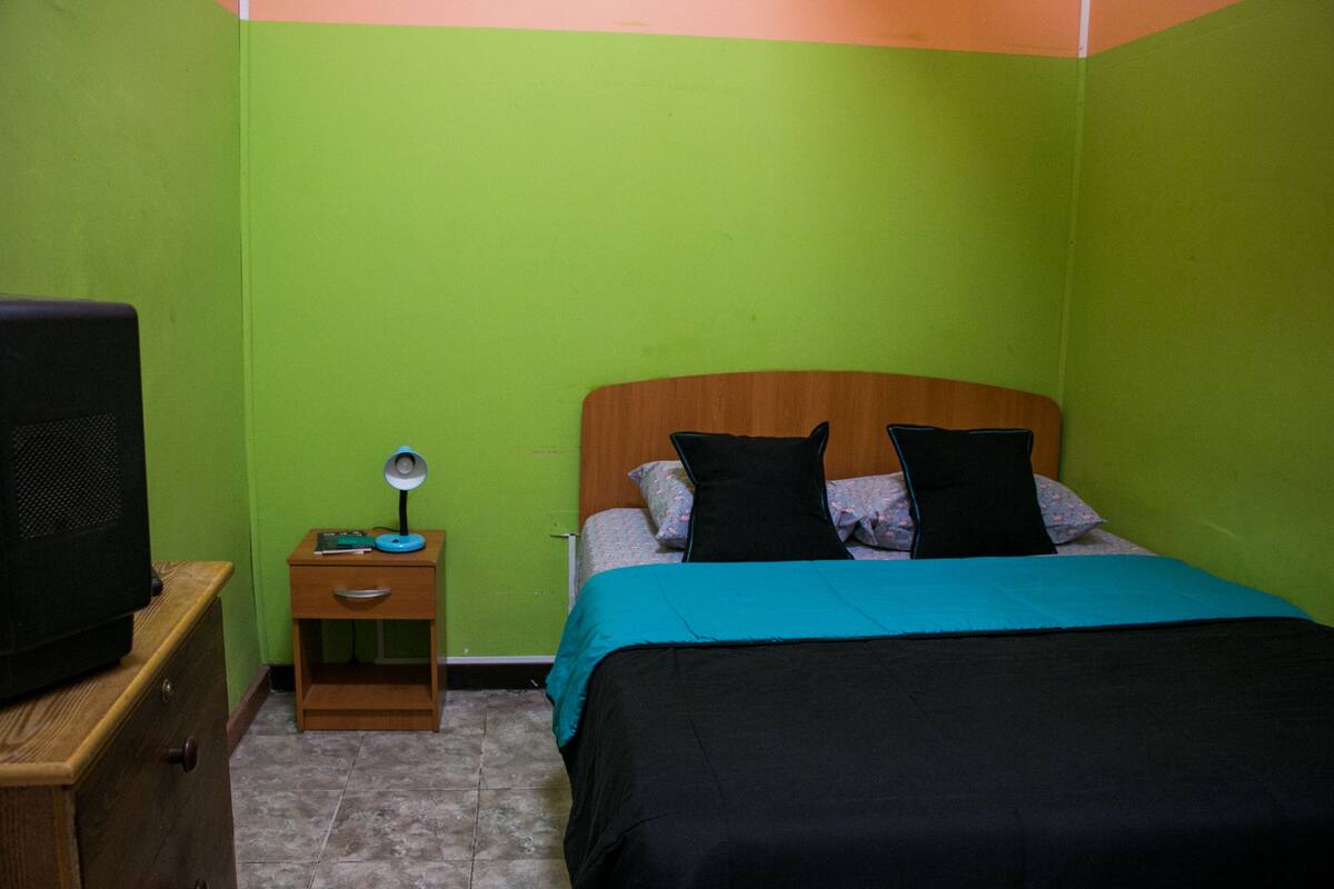 A small, single-bed room with a wooden headboard, a green wall, a small wooden nightstand with a lamp, and a television on a wooden dresser.
