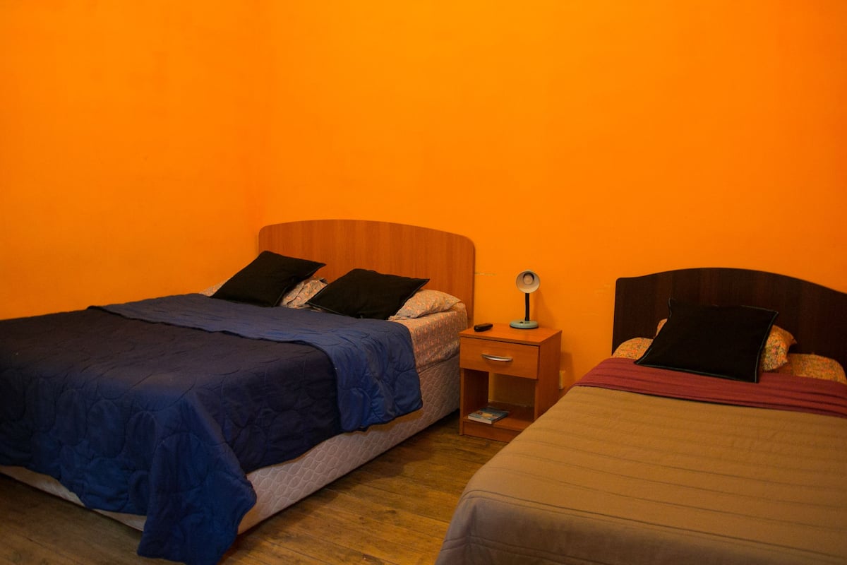 A room with two beds, each with a wooden headboard, a nightstand with a lamp, and a small drawer.