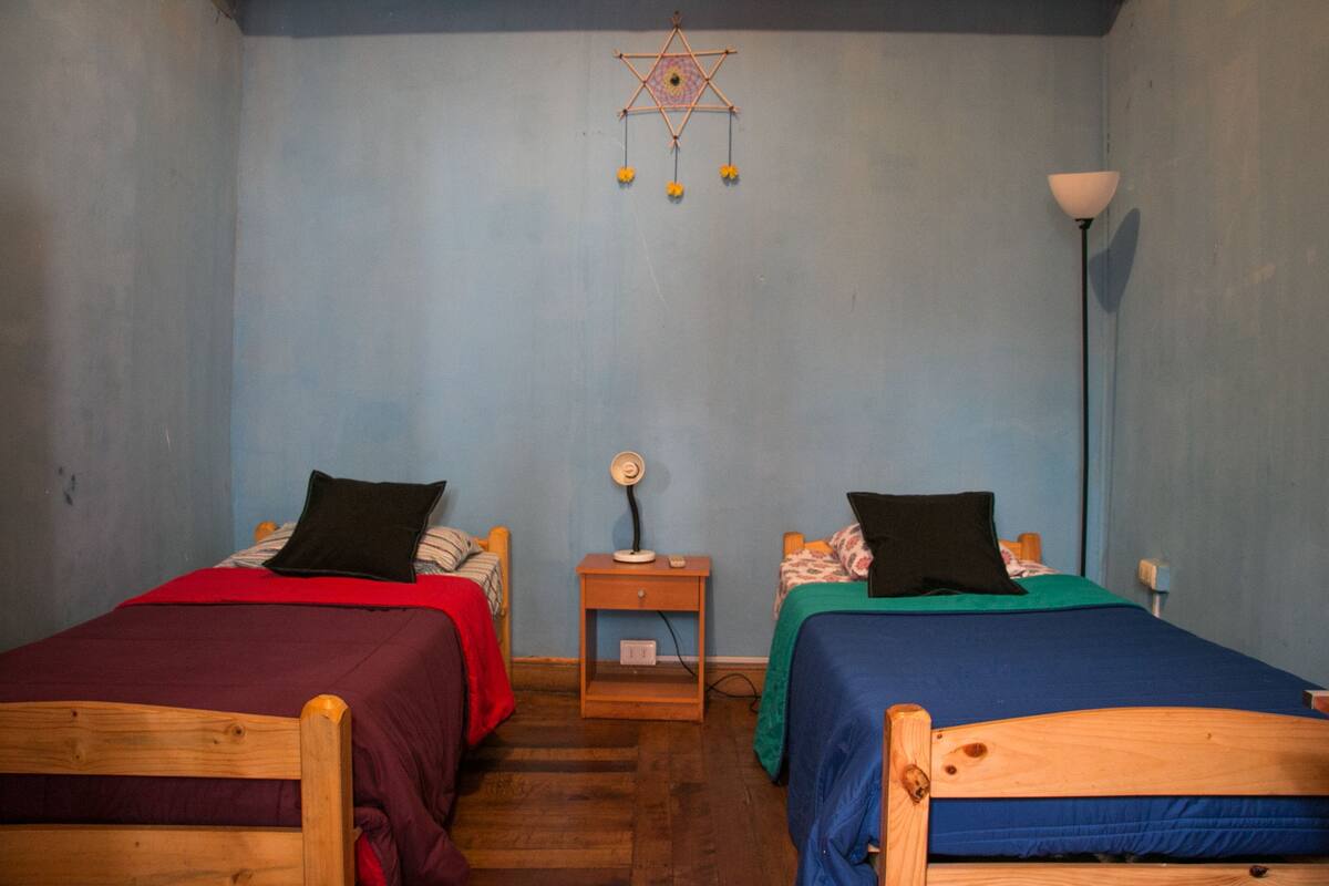 A room with two wooden beds, each with a different colored bedspread and a black pillow. A small wooden nightstand with a lamp is between the beds. The walls are painted blue, and there is a decorative hanging on the wall.