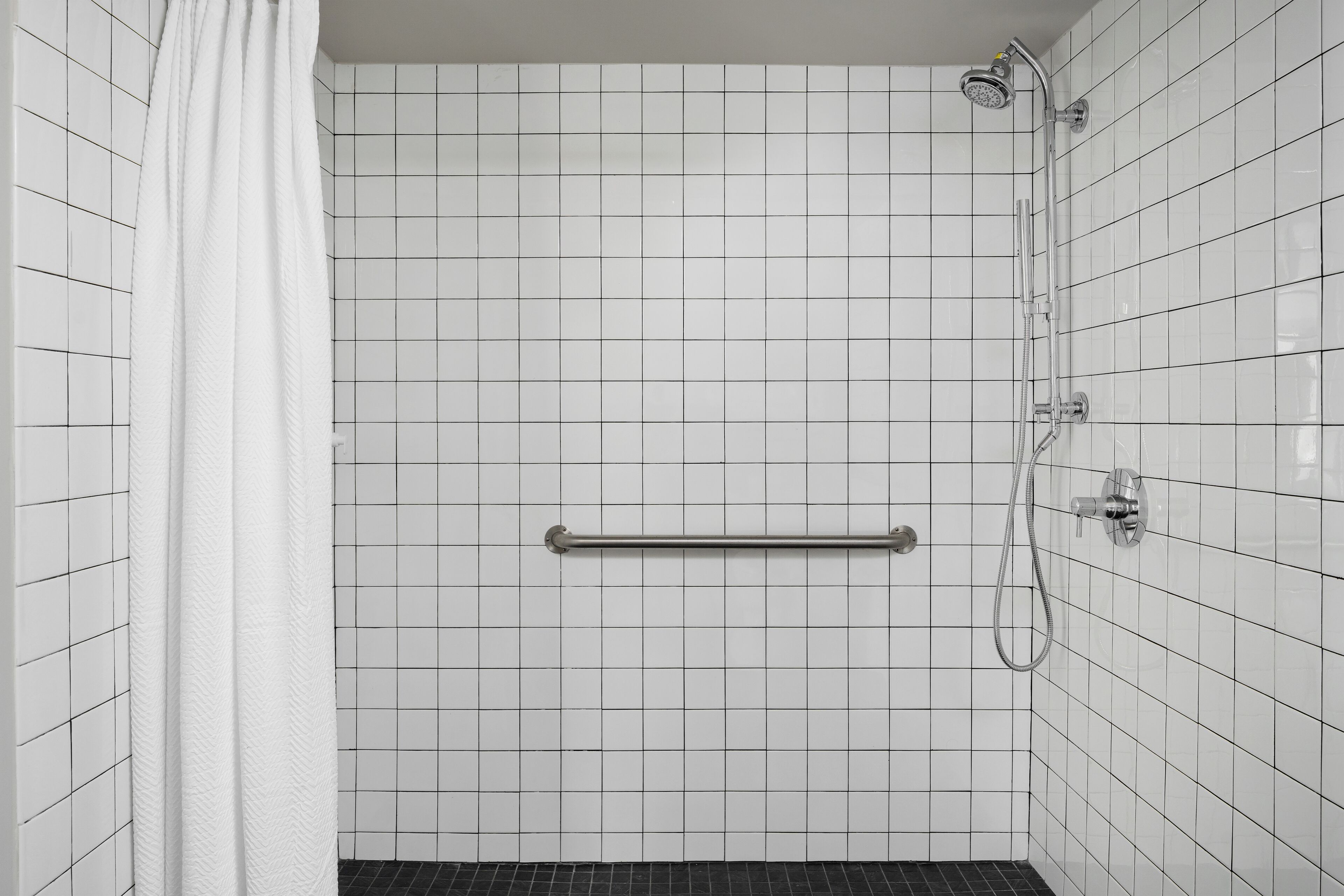 A bathroom with white tiled walls, a shower with a handheld showerhead, and a grab bar.