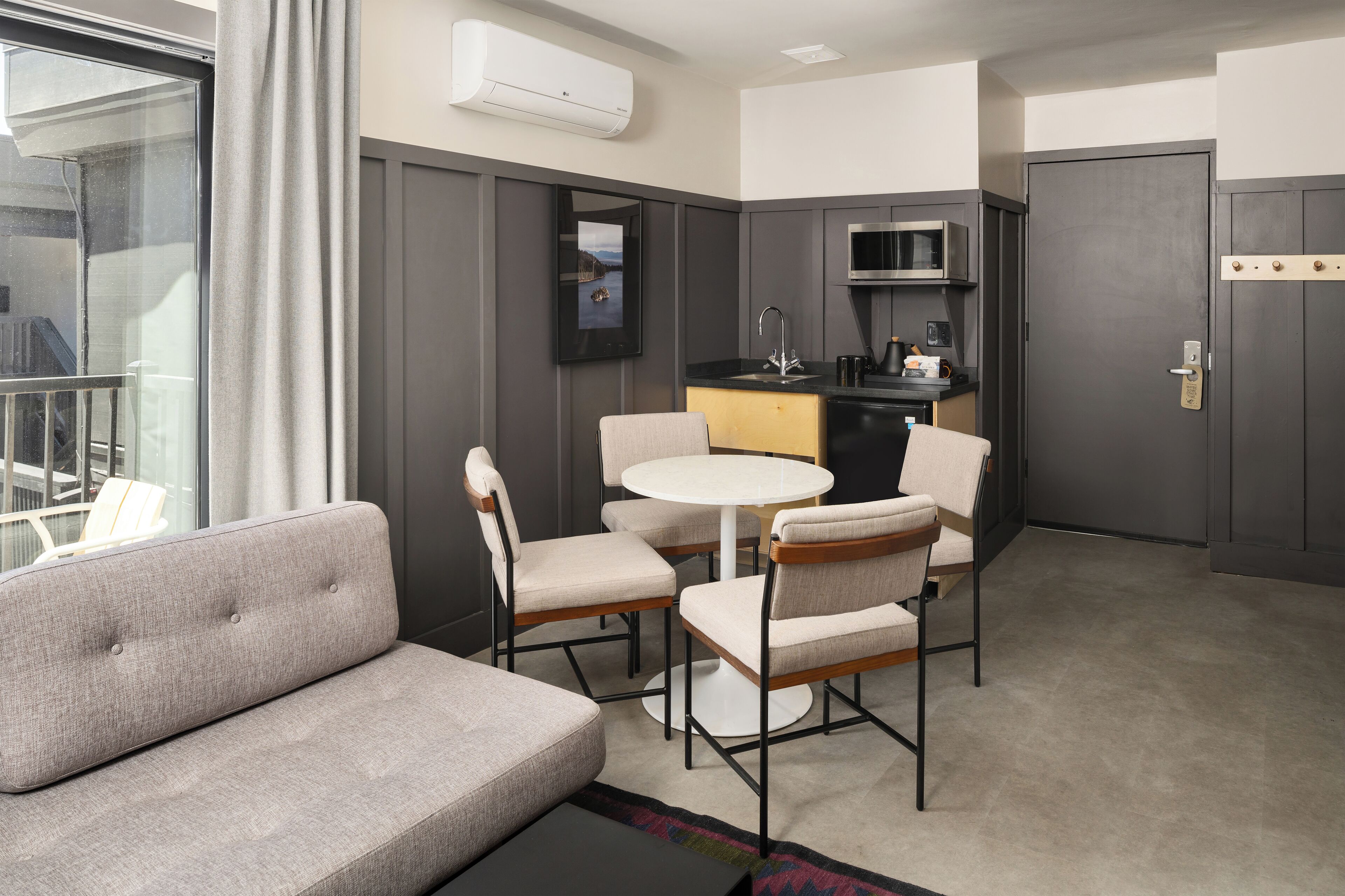 A modern hotel room with a sofa, a round dining table, and four chairs. There is a kitchen area with a microwave and a sink.