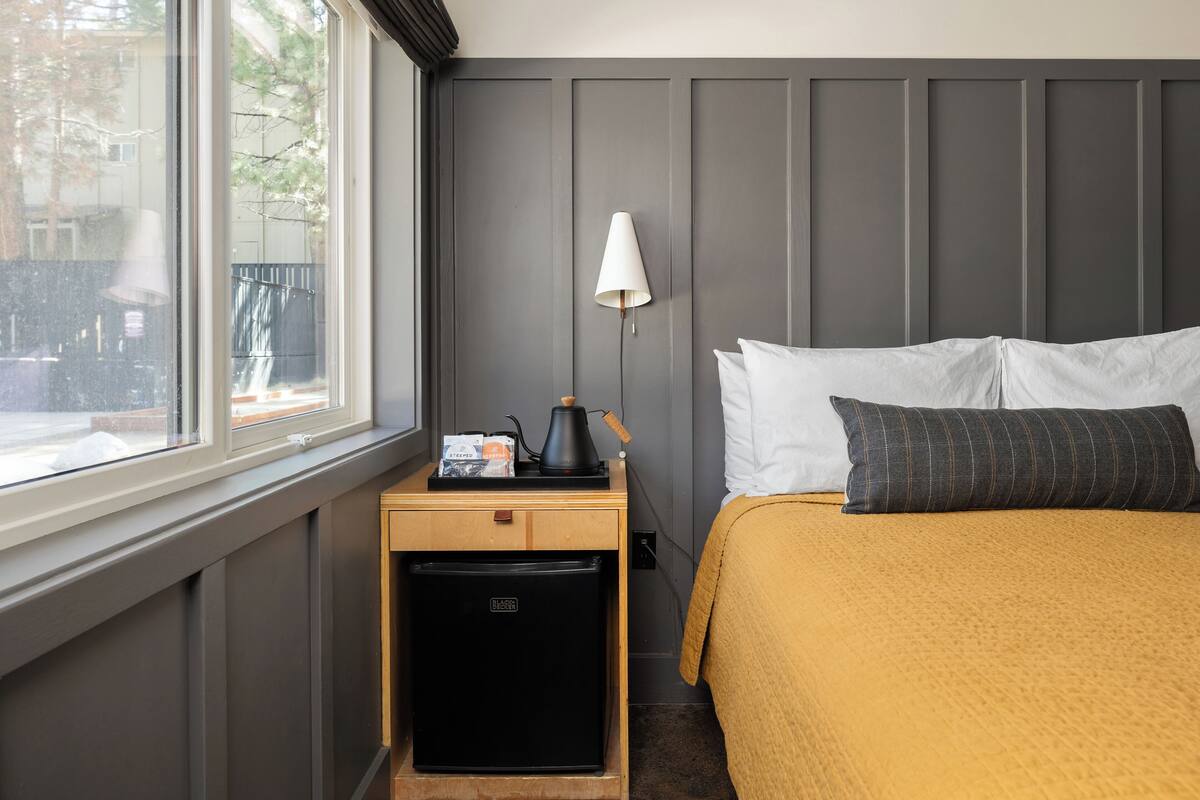 Une chambre d’hôtel équipée d’un lit, d’une table de chevet, d’une lampe, d’un petit réfrigérateur et d’une fenêtre donnant sur l’extérieur.