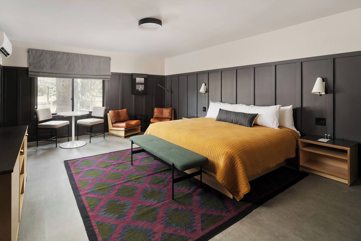 Une chambre d’hôtel moderne avec un grand lit, un bureau, deux chaises, un banc et un tapis à motifs.