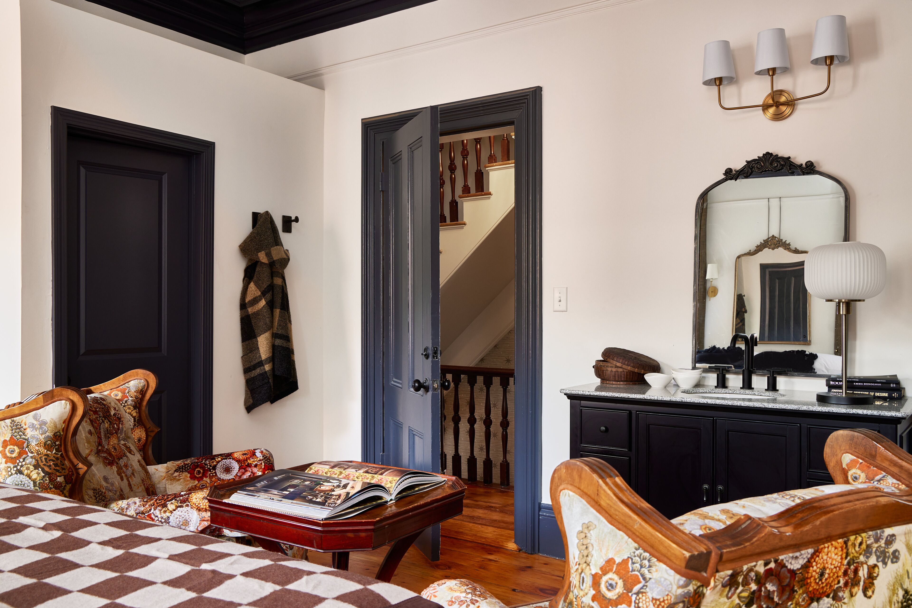 Ein Zimmer mit kariertem Bettbezug, einer Holzbank mit einem Buch, einem an der Wand hängenden Mantel und einer Treppe, die zu einer weiteren Ebene führt.