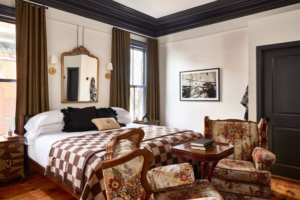 Quarto de hotel com uma cama grande, duas cadeiras, uma mesa, um espelho e um quadro na parede.