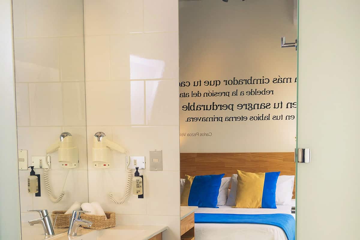 Moderni hotellihuone, jossa on puinen meikkipöytä, sinisinä ja keltaisina sävyinä oleva vuodevaatteisto sekä suuri peili.
