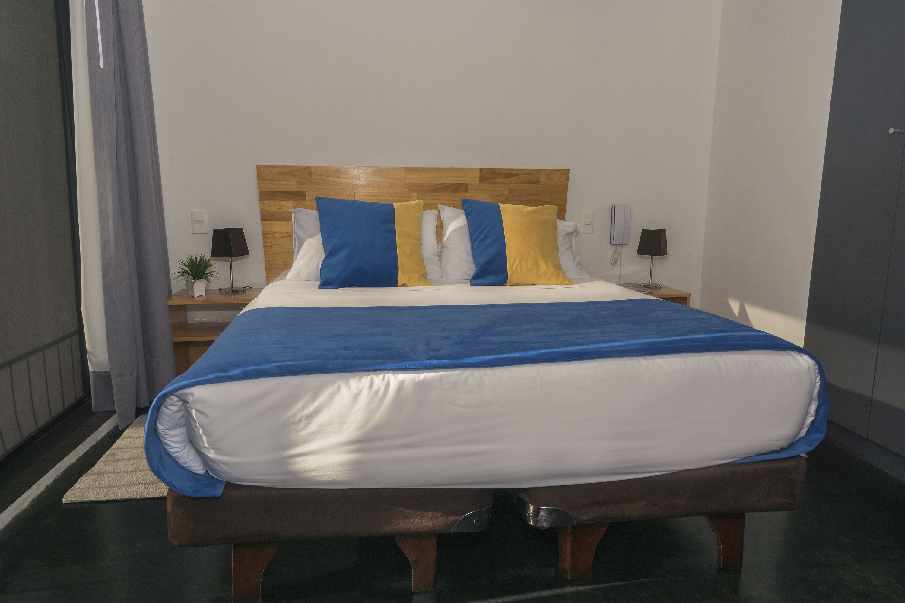 Una cama bien hecha con colcha azul y blanca, cabecera de madera y dos mesitas de noche con lámparas.