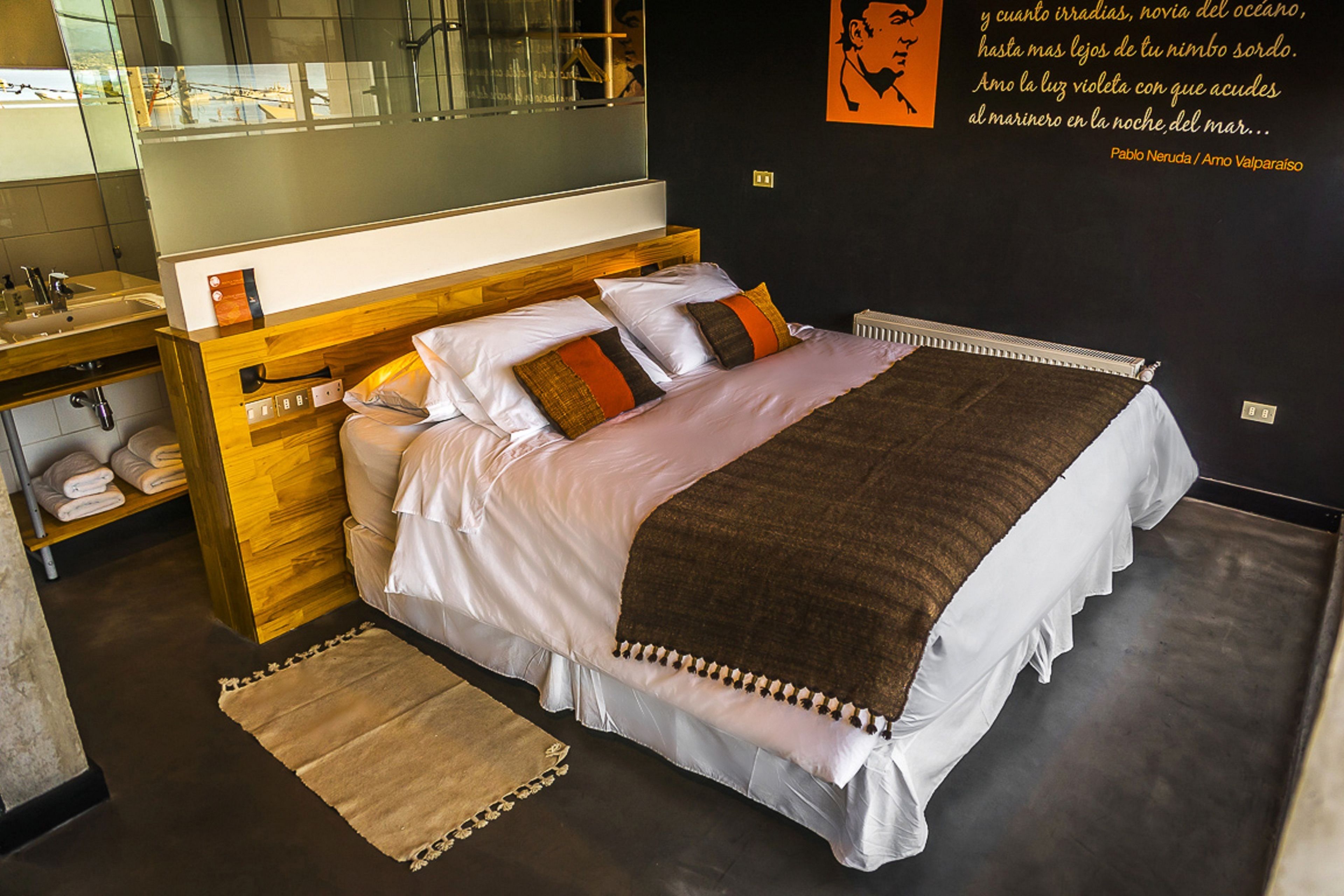 A modern bedroom with a wooden headboard, a large bed with white linens and brown accents, a bedside table with a mirror, and a view of the sea through a large window.