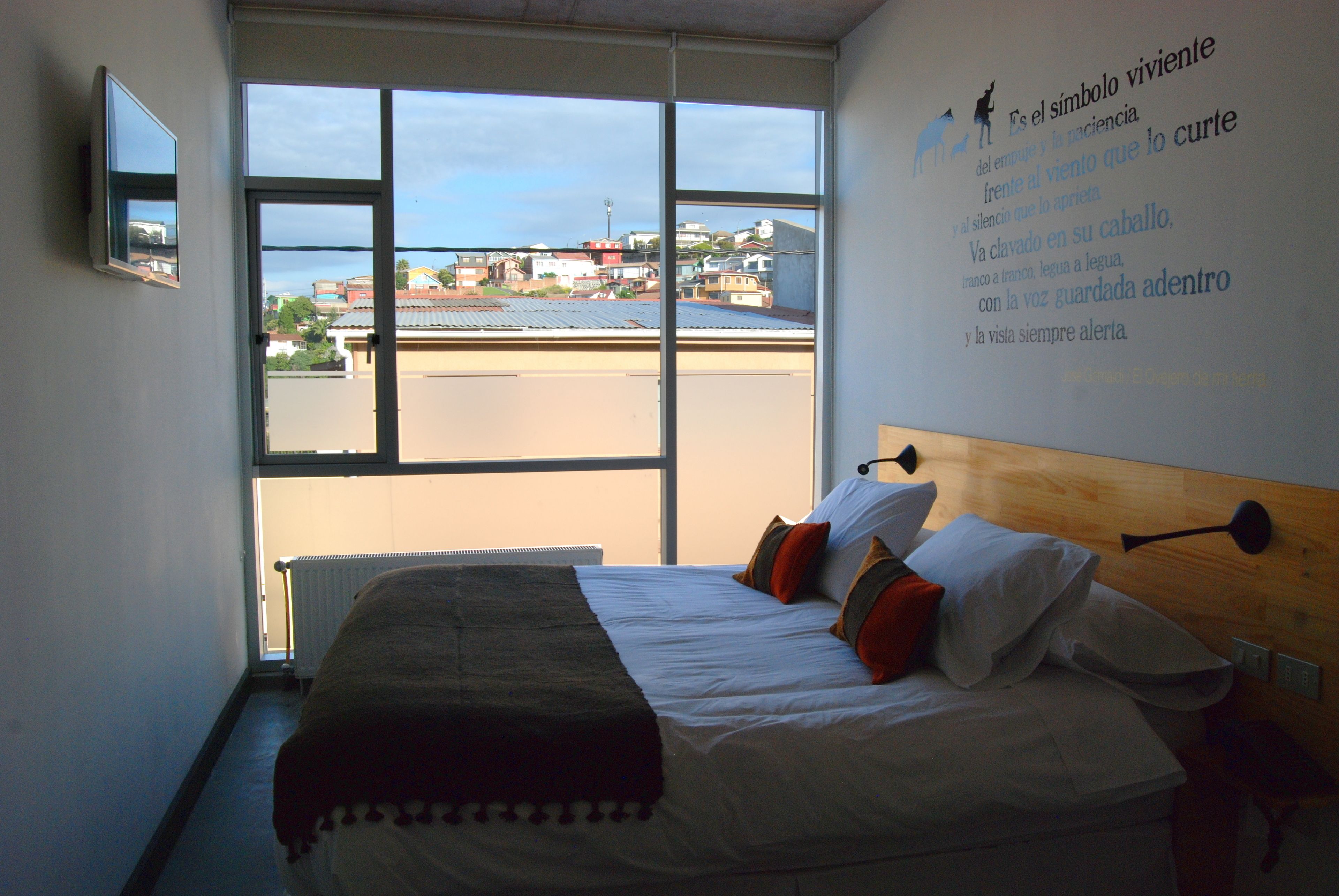 A hotel room with a large window, a bed with pillows, a bedside table, and a wall-mounted TV.