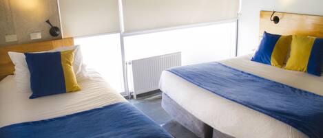 Habitación de hotel con dos camas, cada una con ropa de cama azul y amarilla, cabeceros de madera y un radiador blanco.