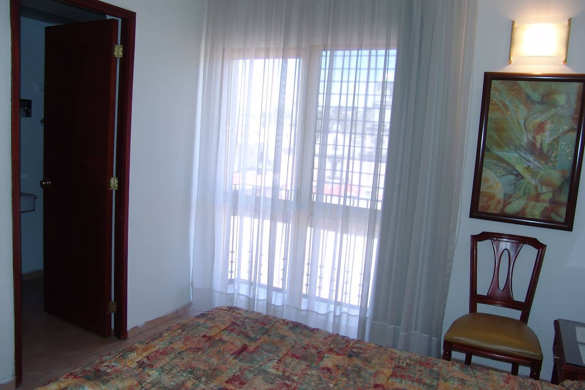 Ein Schlafzimmer mit einem Bett, einem Sessel, einem Bild und einem Fenster mit durchsichtigen Vorhängen.