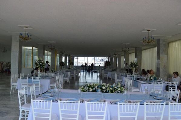 Un salón amplio con filas de mesas y sillas blancas, ventanas grandes y candelabros.