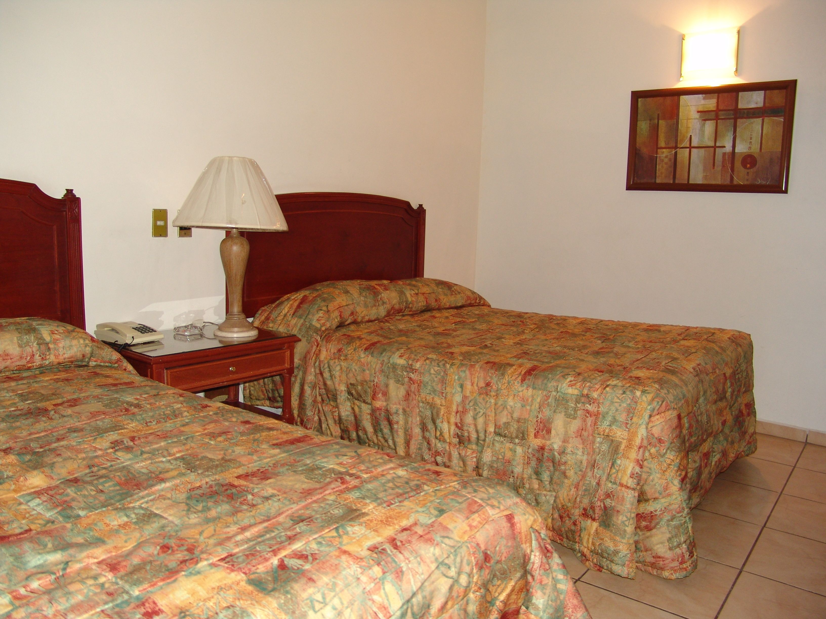 Una habitación de hotel con dos camas, una mesita de noche con lámpara, un teléfono y una obra de arte colgada en la pared.
