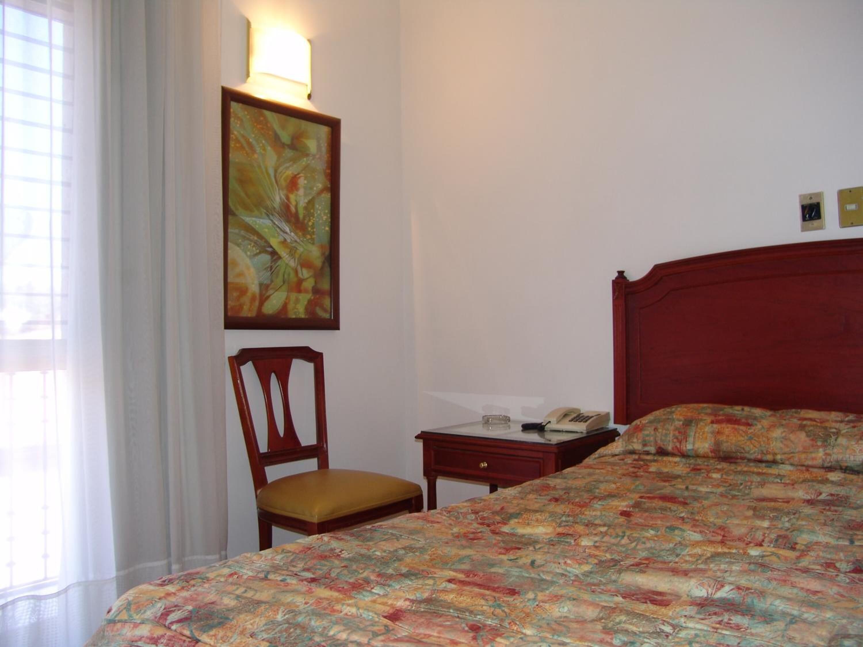 Une chambre d’hôtel avec un lit, une chaise, une table de chevet avec un téléphone et une lampe fixée au mur.