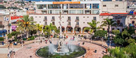 Un hotel con una fuente central, rodeado de palmeras y una plaza adoquinada.