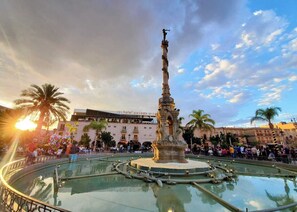 A public square with a central fountain, palm trees, and a hotel named 'hotel posada arc' in the background.