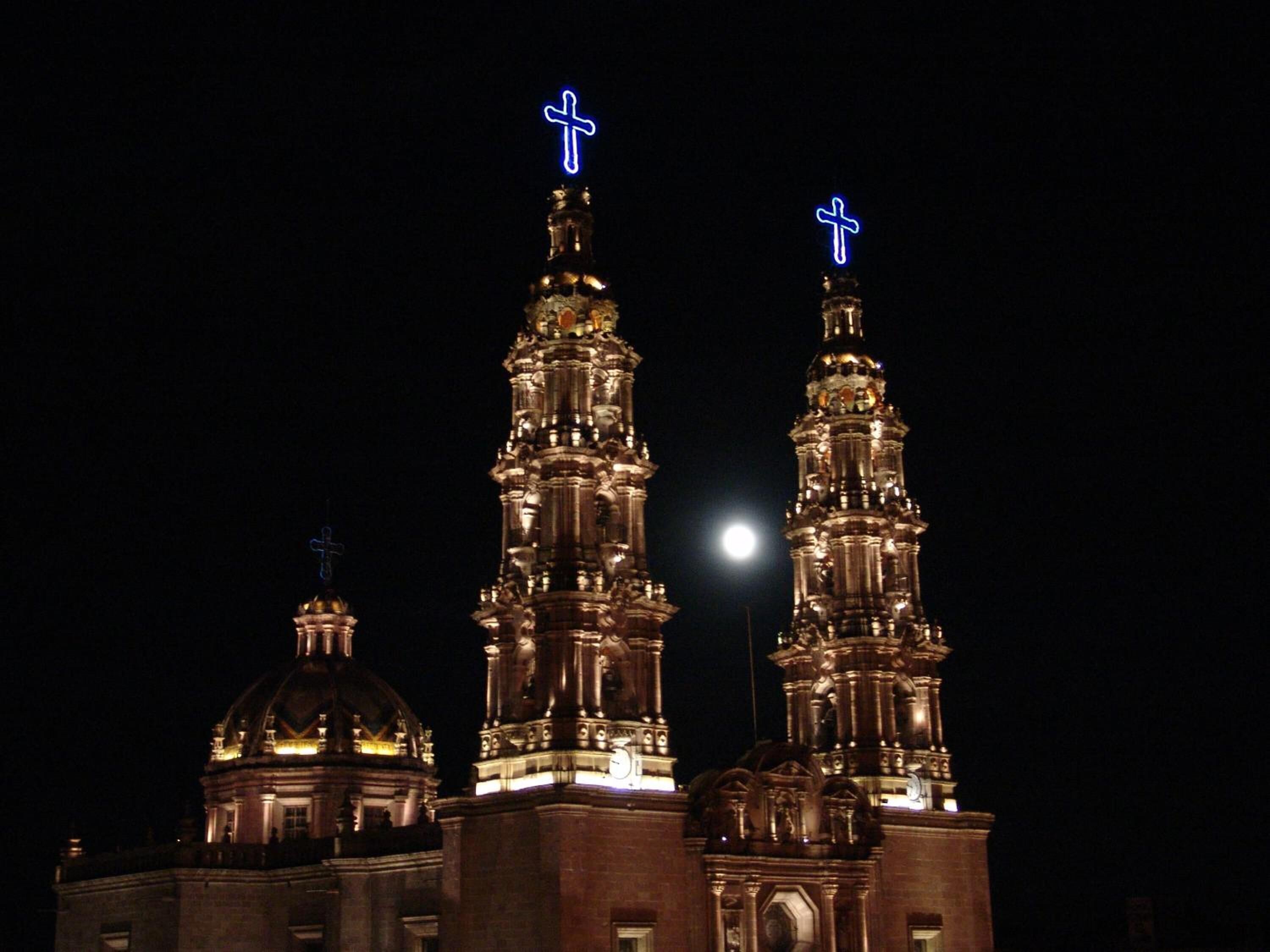 Une église dotée de trois flèches illuminées, chacune surmontée d’une croix, se dressant sous un ciel nocturne où l’on peut apercevoir la lune.