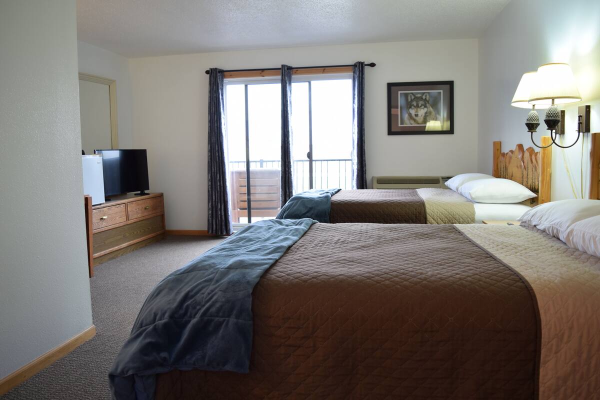 A hotel room with two beds, a television on a dresser, a balcony with curtains, and a framed picture on the wall.