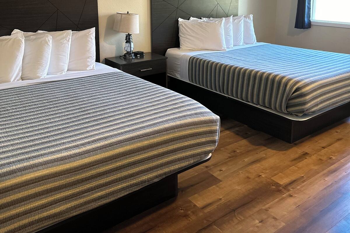 A hotel room with two beds, a nightstand with a lamp, a painting on the wall, and a window with curtains.