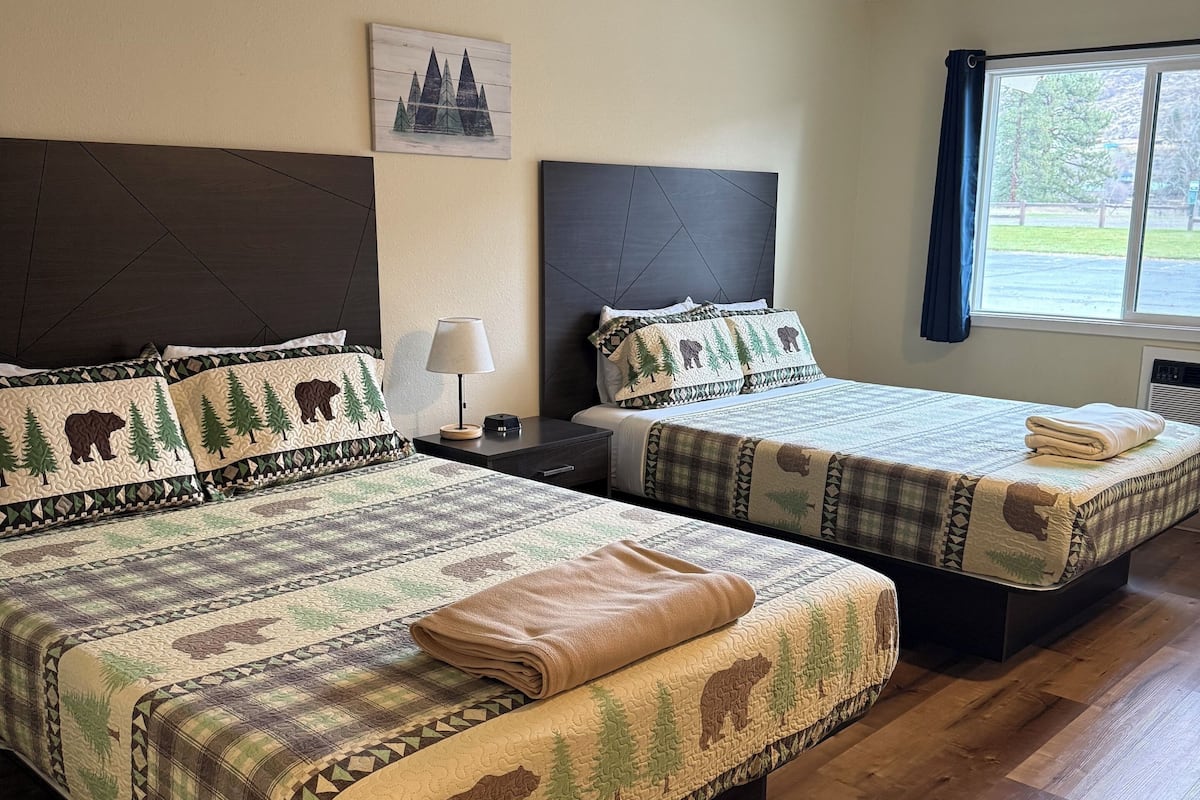 A hotel room with two beds, each with a forest-themed bedspread and headboard. There is a window with a view of the outdoors, a nightstand with a lamp, and a wall-mounted painting.