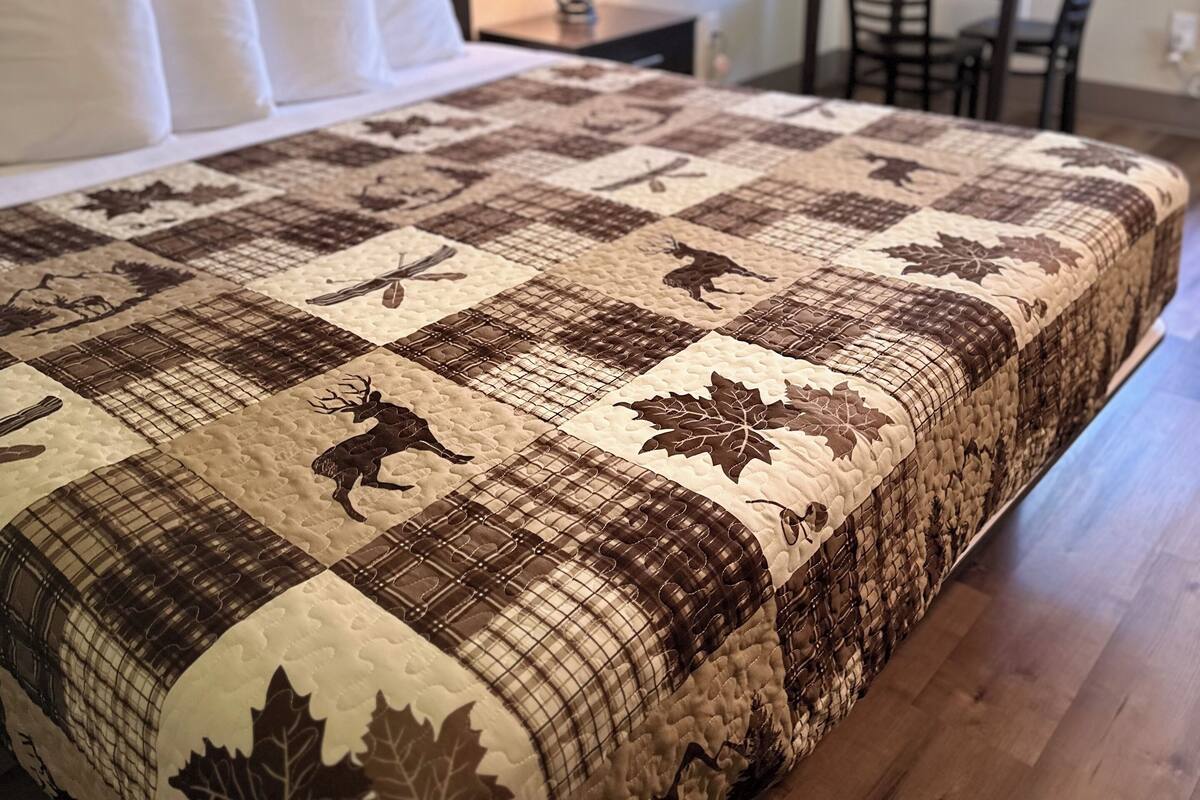A neatly made bed with a quilt featuring animal and leaf patterns, a bedside lamp, a small table, and a window with a view of greenery.