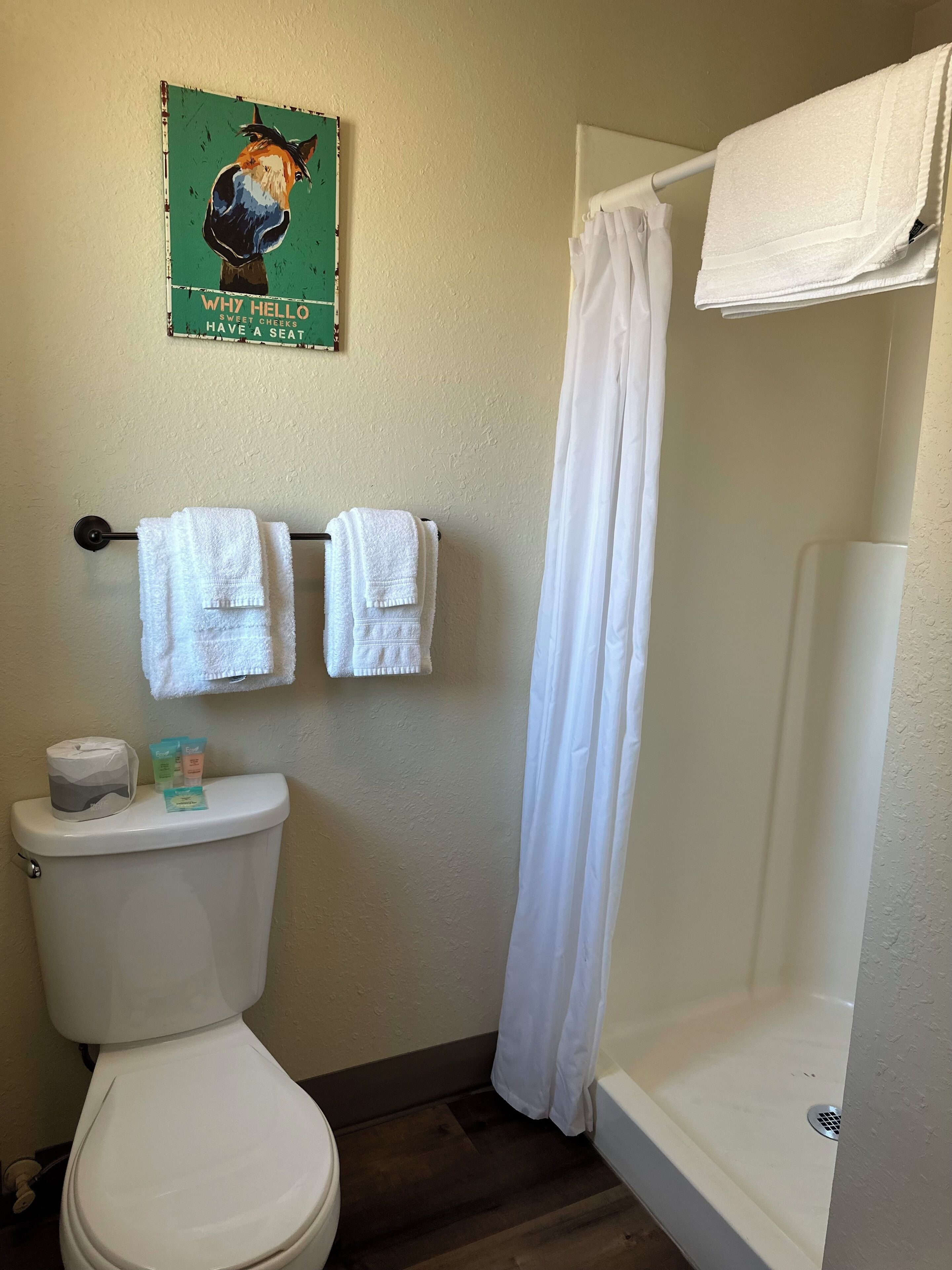 A bathroom with a toilet, two towels on a rack, a shower curtain, and a sign on the wall.