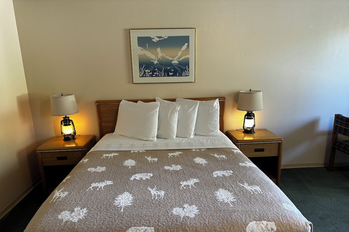 A hotel room with a bed, two bedside lamps, a framed picture on the wall, and a chair in the corner.