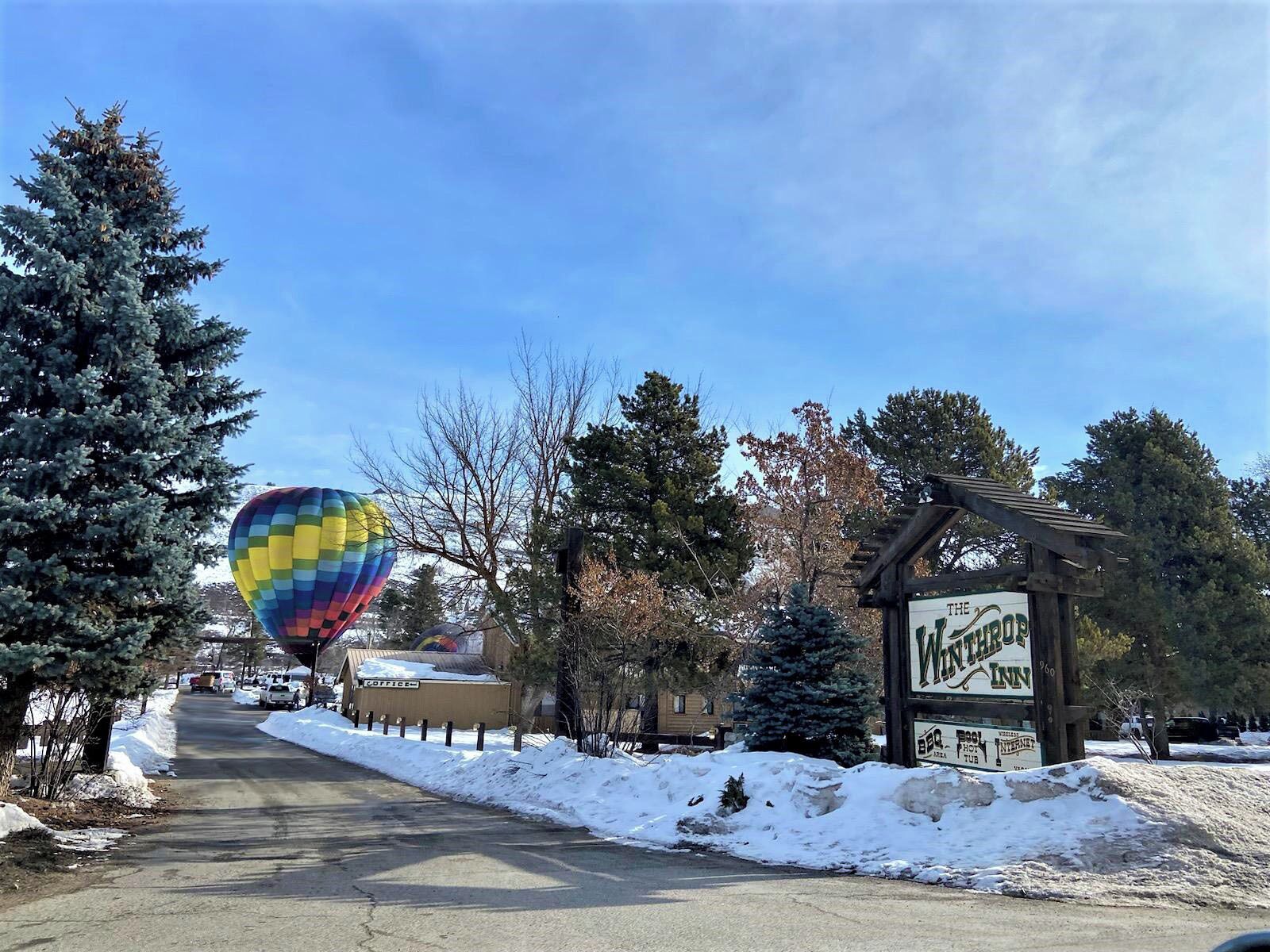 A snow-covered road with a hot air balloon in the distance, a sign for 'The Wilderness Inn', and snowbanks along the roadside.
