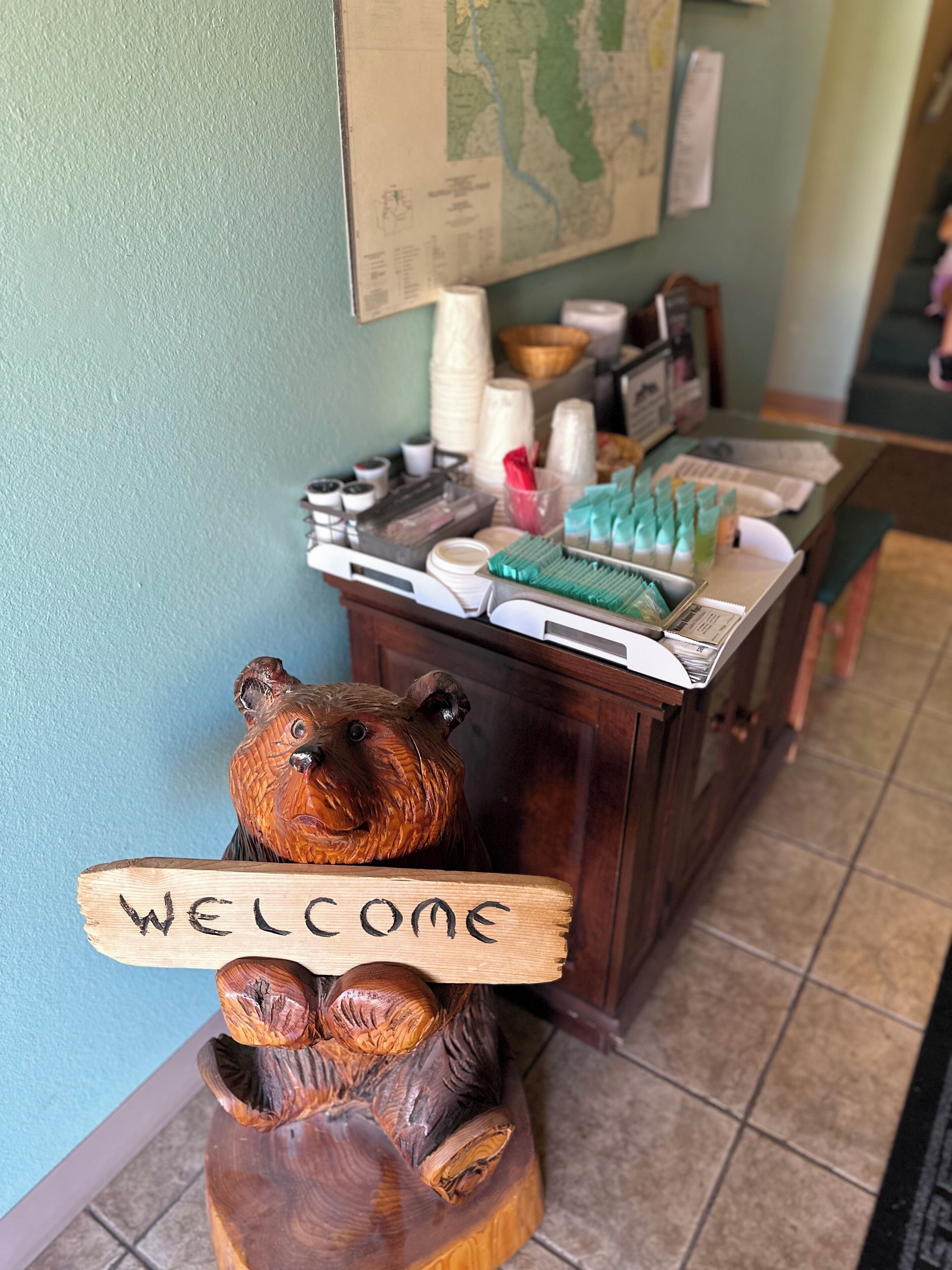 Une sculpture en bois représentant un ours tenant une pancarte sur laquelle est écrit « Bienvenue », un bureau avec divers objets et une carte accrochée au mur.