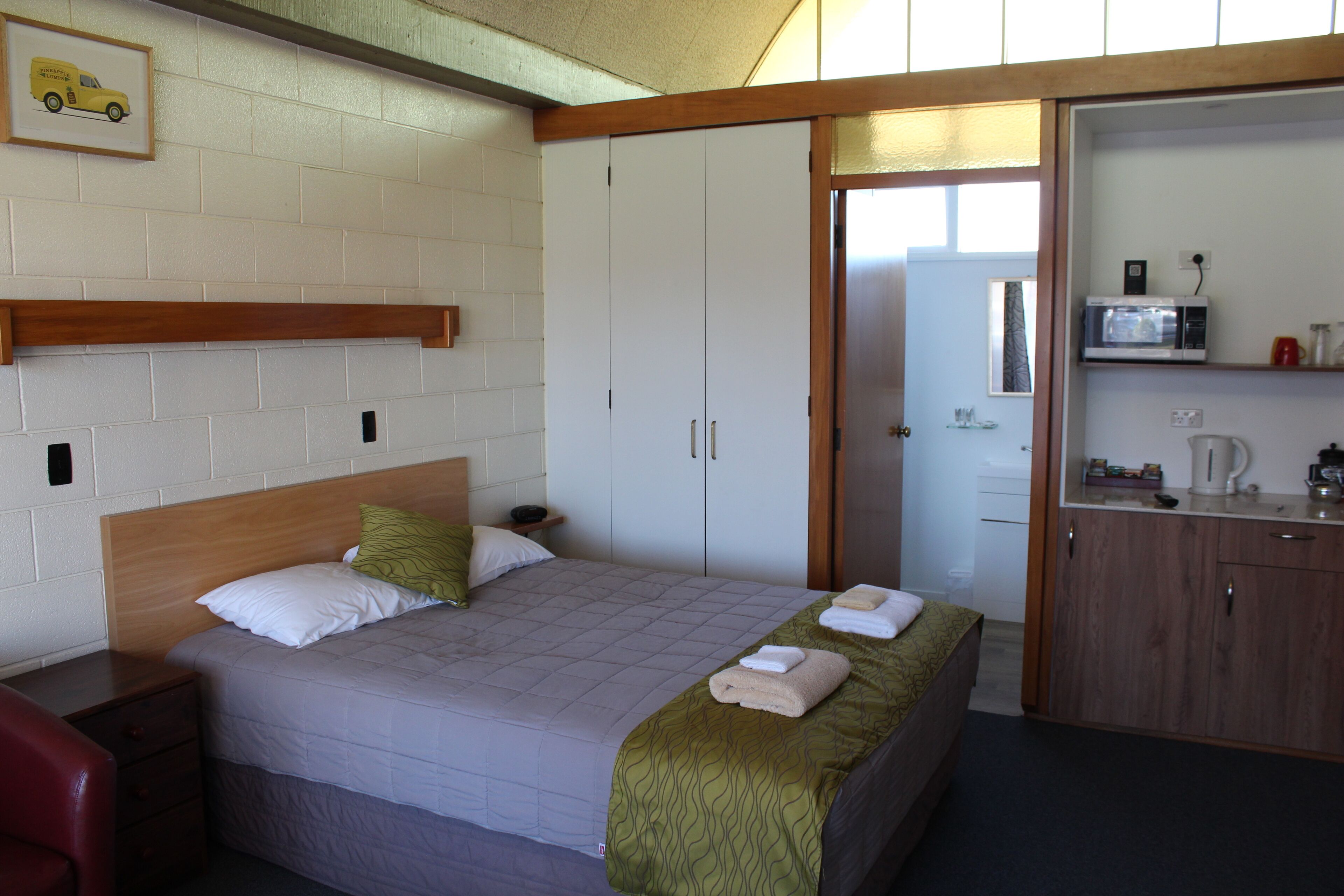 A hotel room with a bed, a wooden headboard, a bedside table, a red chair, a wardrobe, and a kitchenette with a microwave.