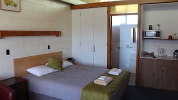 A hotel room with a bed, a wooden headboard, a bedside table, a red chair, a wardrobe, and a kitchenette with a microwave.
