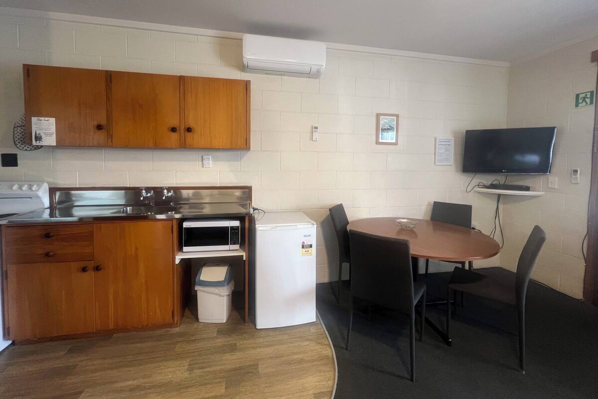 A compact kitchen with wooden cabinets, a microwave, a stove, and a small refrigerator. There is a dining table with chairs and a flat-screen TV mounted on the wall.