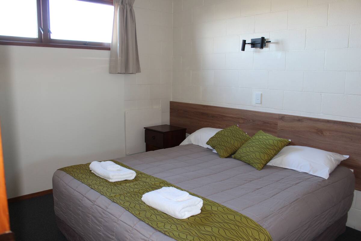 A neatly made bed with a green bedspread, two pillows, and two folded towels.