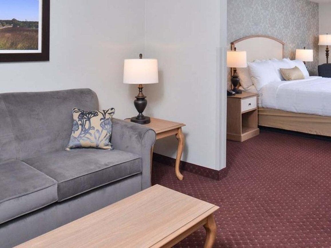 Quarto de hotel com um sofá cinzento, uma mesa de madeira, duas luminárias de cabeceira, uma cama com roupa de cama branca e um quadro na parede.