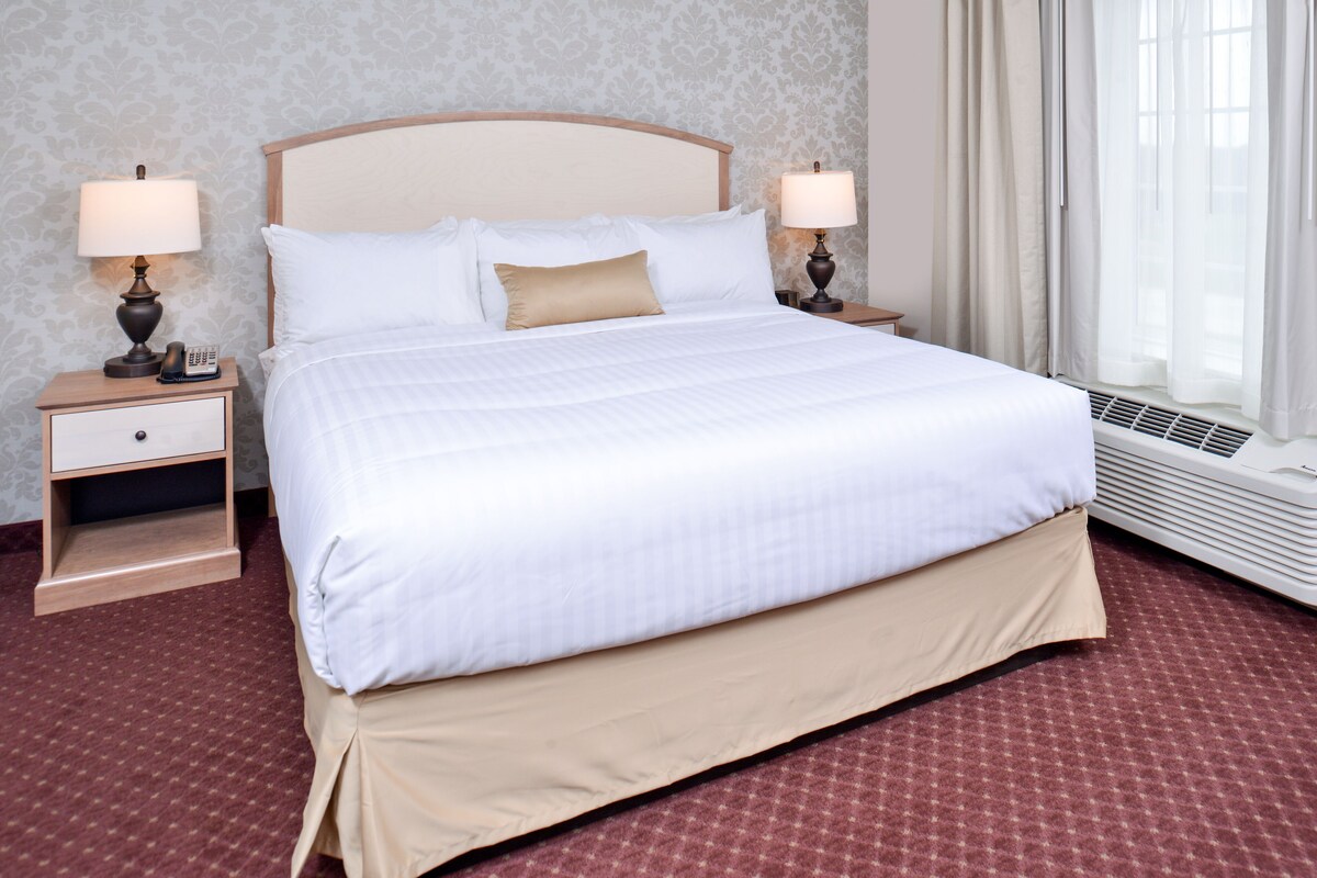 Een netjes opgemaakt bed met witte lakens en een beige onderrok, twee nachtkastjes met lampen, een kamerbreed tapijt met dessin, en een raam met dunne gordijnen.