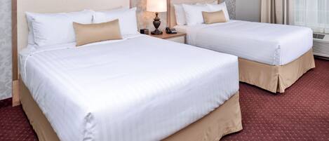 A hotel room with two beds, each with white bedding and beige accents. There is a nightstand with a lamp and a telephone between the beds. The room has a patterned wallpaper and a red carpet.