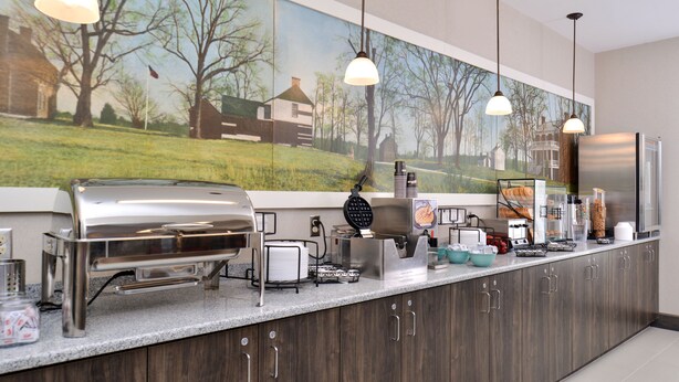 A breakfast bar with a stainless steel waffle maker, coffee station, and various breakfast items. - Appomattox Inn and Suites (Appomattox)