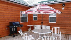 Una terraza de madera con una parrilla, una mesa redonda con sillas y un paraguas a rayas.
