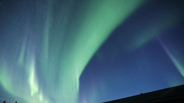 Un edificio con ventana y puerta visibles, bajo un cielo nocturno lleno de estrellas y la aurora boreal.
