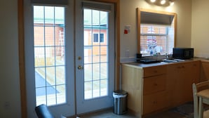 Una cocina pequeña con armarios de madera, un lavamanos y un microondas. Hay dos ventanas que dan a una terraza de madera.