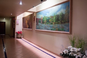 A hallway with two large paintings, a potted plant with red flowers, and a decorative arrangement of white flowers and green foliage. - Hotel Real Santa Fe (San Juan de los Lagos)
