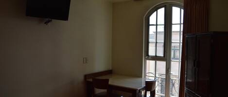 A room with a bed, a desk, a chair, a television mounted on the wall, and a balcony with a view of the outside.
