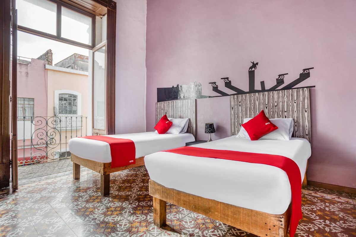 Hotellihuoneessa on kaksi sänkyä, joissa on punainen peitto, valkoiset lakanat ja punaiset tyynyt. Huoneessa on kuvioitu lattia ja suuri ikkuna, josta on näkymä ulkona oleviin rakennuksiin.