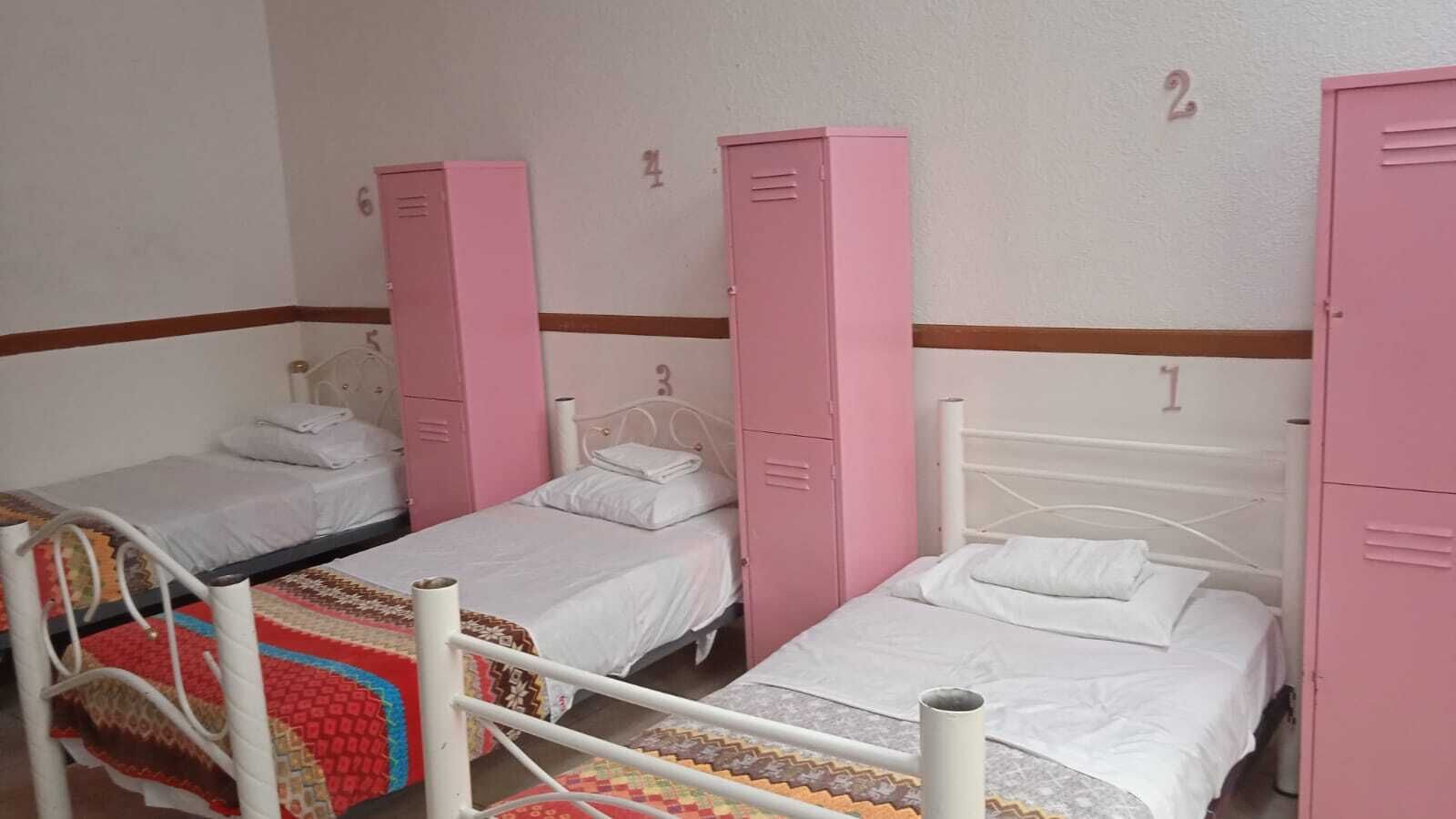 Habitación tipo dormitorio con tres camas individuales, cada una con colchón y almohadas blancos. Hay taquillas rosas con los números 1, 2 y 3 en la pared.
