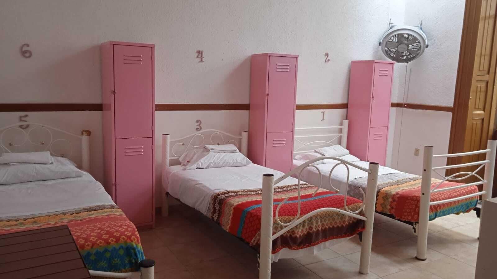 A dormitory room with three beds, each with a different patterned bedspread. There are pink lockers on the wall, and a ceiling fan is visible.